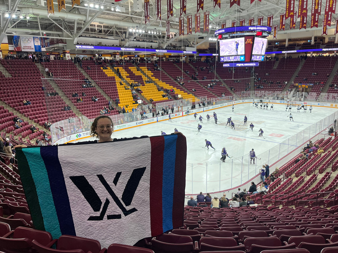 Woman holding a quilt stands overlooking the ice at a collegiate hockey rink. Players warm up on the ice behind her, with spectators trickling into the maroon and gold seats.